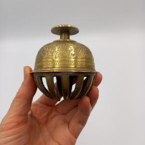 May include: A vintage brass bell with a floral pattern etched around the top. The bell has a rounded shape with vertical slots at the bottom. The bell is held in a hand against a white background.