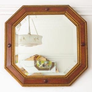 May include: An octagonal wall mirror featuring a warm brown wooden frame with gold-toned beaded detailing along the inner and outer edges. The frame is accented with four circular wooden studs positioned at the top, bottom, left, and right sides.