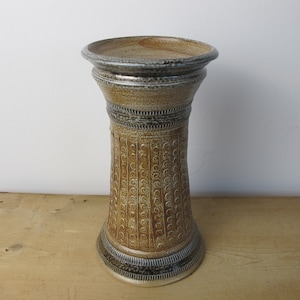 May include: A tall, cylindrical ceramic vase with a wide, flared base and a slightly wider top. The vase is primarily tan with decorative vertical patterns and bands of gray and black near the top and bottom. It sits on a wooden surface.