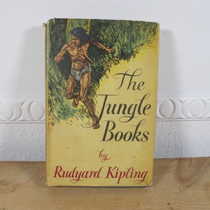 Peut inclure: Un livre relié vintage avec une couverture jaune et une illustration colorée d'un homme courant dans la jungle. Le titre "The Jungle Books" est écrit en cursive avec l'auteur "Rudyard Kipling" en dessous.