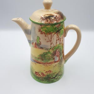 May include: A cream-colored ceramic coffee pot with a green and brown landscape scene painted on the side. The pot has a lid and a handle.