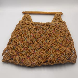 Puede incluir: Un bolso tote tejido en crochet color marrón con un asa de madera. El bolso presenta una superposición de crochet con patrón de diamantes, que revela un forro de tela floral con flores rojas, amarillas y verdes. El asa es de madera cilíndrica marrón claro.