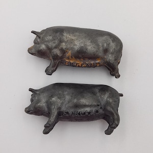 Könnte beinhalten: Zwei antike Metall-Schweinefiguren mit einem verwitterten, dunklen Finish. Das Wort "ENGLAND" ist auf der Seite eines Schweins eingeprägt, und das andere Schwein hat das Wort "ENGLAND" auf seiner Seite eingeprägt. Die Schweine sind auf einer weißen Oberfläche positioniert.