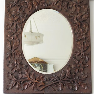 May include: A rectangular wall mirror featuring a dark brown wooden frame with intricate hand-carved floral and leaf motifs surrounding a central oval glass pane. The wood carving displays detailed petals and foliage patterns throughout the border.