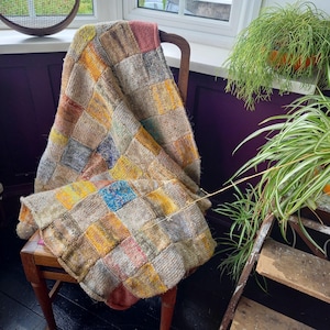 May include: A handmade patchwork blanket draped over a wooden chair. The blanket features a variety of colorful squares in shades of yellow, blue, pink, and gray. The blanket is made of a textured material, possibly wool or a similar fiber.