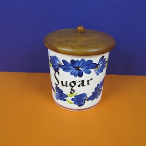 May include: A white ceramic sugar container with a wooden lid. The container is decorated with blue floral designs and the word "Sugar" in black script. The lid has a small wooden knob.
