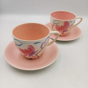 May include: Two vintage teacups and saucers. The cups have a light pink interior and are decorated with pink and purple floral designs on a cream background. The saucers are light pink.