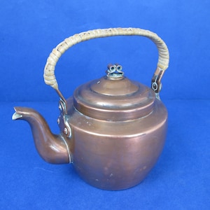 Op de afbeelding: Een koperen theepot met een gevlochten rieten handvat. De theepot heeft een rond lichaam en een deksel met een decoratieve knop.