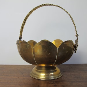 May include: A vintage brass basket with a petal-shaped bowl and a twisted handle. The basket sits on a round base and features decorative floral accents. The brass has a warm, golden hue.