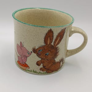 May include: Beige ceramic mug with a green rim and handle. The mug features a whimsical illustration of a brown rabbit and a pink mouse. The rabbit and mouse are facing each other. The mug is decorated with grass and a mushroom.