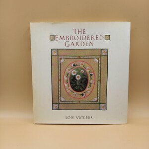 Könnte beinhalten: Ein Buch mit dem Titel "The Embroidered Garden" von Lois Vickers. Das Cover zeigt ein zentrales quadratisches Bild von gestickten Gänseblümchen in einem dekorativen Rahmen. Das Buch hat einen cremefarbenen Hintergrund.