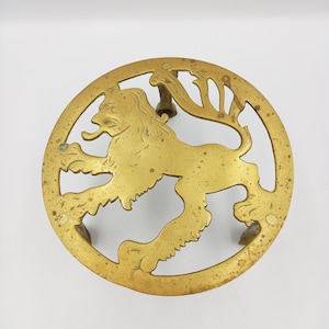 Op de afbeelding: Een ronde messing trivet met een leeuwenontwerp. De leeuw staat met zijn staart omhoog gekruld. De trivet heeft drie poten.