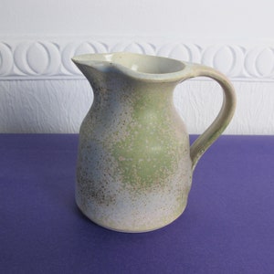 Puede incluir: Una pequeña jarra de cerámica moteada con un esmalte verde claro y blanco. La jarra tiene un cuerpo redondeado y un asa.