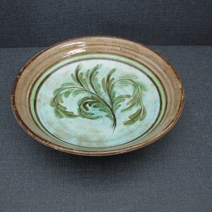 Puede incluir: Un cuenco de cerámica para servir con un diseño pintado a mano. El cuenco presenta un interior azul claro con un patrón de hojas verdes y marrones. El borde es marrón con un acento verde. Una pieza decorativa para la cocina o el comedor.