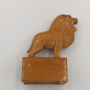 Op de afbeelding: Een houten leeuwenbeeldje, gesneden in een staande positie met een open mond. De leeuw heeft een gedetailleerde manen en is gemonteerd op een rechthoekige basis. De kleur is lichtbruin. De achtergrond is effen wit.