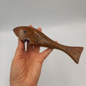 May include: A brown wooden bottle opener shaped like a fish. The fish features carved crescent-shaped scales along its body, a small dark eye, and a metal bottle opening mechanism integrated into the mouth area of the wooden fish figure.