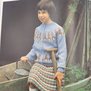 May include: A light blue knit cardigan featuring a repeating brown and white rabbit pattern around the yoke and hem, secured with light blue buttons. The garment is paired with a skirt featuring horizontal stripes in shades of brown, green, and white.