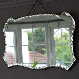 Pode incluir: Um espelho antigo e decorativo com um design de borda exclusiva e ondulada. O espelho retangular tem uma parte superior e laterais onduladas e é suspenso por uma corrente. O vidro reflete uma janela com vista para árvores e uma casa.