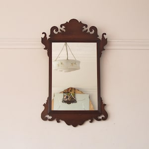 Puede incluir: Un espejo de pared rectangular con un marco de madera de color marrón oscuro que presenta tallas ornamentadas y curvas en la parte superior e inferior. La superficie de cristal refleja una lámpara colgante con pantalla blanca y otro espejo.