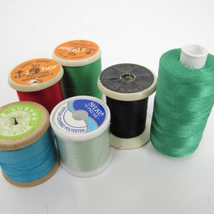 May include: Six spools of thread in various colors, including red, green, blue, black, and white. The spools are made of wood and plastic and have different labels, including "Anchor", "Sylko", "Lustre", and "Clark & Co."