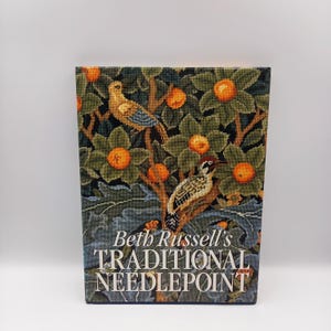 Op de afbeelding: Een boek getiteld "Beth Russell's Traditional Needlepoint" met een kleurrijk borduurwerkontwerp op de omslag met vogels en fruit op een boom.
