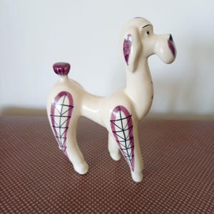 May include: A cream-coloured ceramic poodle figurine with purple and black detailing. The poodle features purple markings on its ears and legs, and a small purple tail. The dog is standing on all fours, a decorative item.