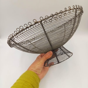 May include: A grey wire pedestal bowl featuring a circular base and a wide, open-work basin. The rim is decorated with a repeating series of small, interconnected wire loops, creating a textured edge around the entire circumference of the bowl.