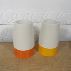 May include: Two white ceramic egg cups with orange and yellow bands. The egg cups are on a wooden surface.