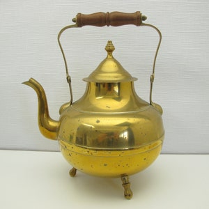 May include: A vintage brass teapot with a rounded body, a curved spout, and a wooden handle. The teapot has a lid with a decorative finial and rests on three small feet. The brass has a warm, golden color.
