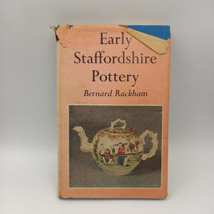 May include: A hardcover book titled Early Staffordshire Pottery by Bernard Rackham with a peach-colored dust jacket. The cover features a central rectangular image of a cream-colored teapot with colorful painted figures and a floral design.