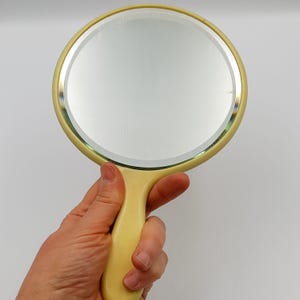Könnte beinhalten: Eine Hand hält einen gelben, handlichen Spiegel aus Kunststoff mit einer runden, klaren Glasfläche.
