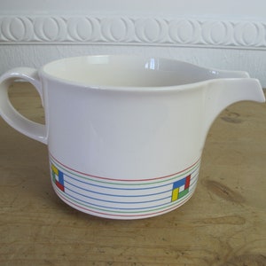 Puede incluir: Jarro de cerámica blanca con asa y pico. El jarro tiene un diseño geométrico colorido alrededor del borde inferior con cuadrados y líneas rojas, amarillas, azules y verdes.