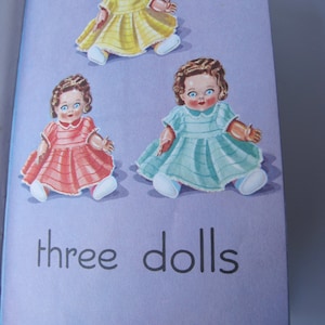 May include: Three dolls wearing dresses. The doll on the left is wearing a pink dress, the doll in the middle is wearing a yellow dress, and the doll on the right is wearing a blue dress. The text "three dolls" is printed below the dolls.
