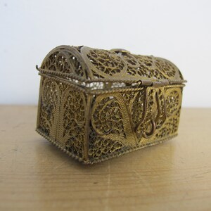 May include: A small, ornate, gold-coloured metal box with a hinged lid. The box is made of filigree, a delicate, intricate design of interwoven metal threads.