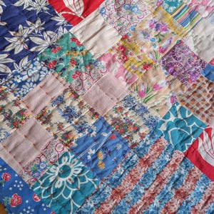 May include: A patchwork quilt constructed from various fabric squares featuring diverse patterns including floral prints, geometric shapes, paisley, and stripes in a vibrant colour palette of red, blue, pink, yellow, white, and teal with visible stitching.