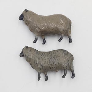 Peut inclure: Deux figurines miniatures de moutons. Les moutons sont peints dans des tons de gris et de beige, avec des visages et des pattes noirs. Les figurines sont détaillées avec de la laine texturée et sont posées sur une surface blanche.