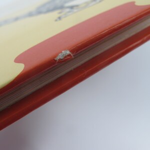 May include: Close-up of a book's corner, showcasing a red cover with a yellow and red design. The book's pages are visible, and there's a small area of damage on the cover's edge. The background is plain white.