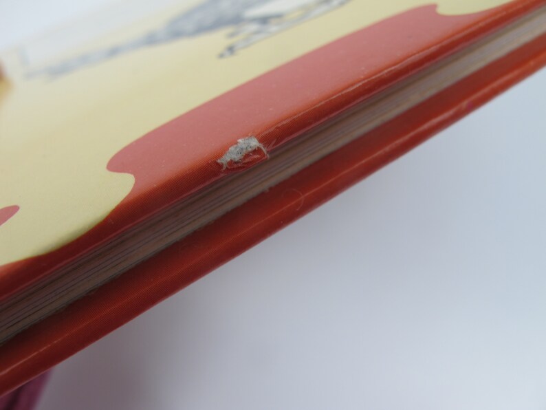 May include: Close-up of a book's corner, showcasing a red cover with a yellow and red design. The book's pages are visible, and there's a small area of damage on the cover's edge. The background is plain white.