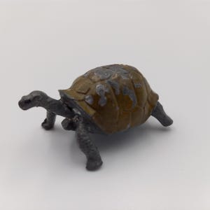 May include: A small, weathered toy tortoise with a brown and grey shell and grey legs and head. The tortoise is positioned on a white surface, showcasing its detailed features and aged appearance. This figurine is a collectible item.
