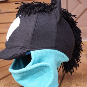 Puede incluir: Un gorro de caballo de lana negra y turquesa con una crin negra y una mancha blanca en la frente. El gorro tiene una visera y una m&aacute;scara facial.