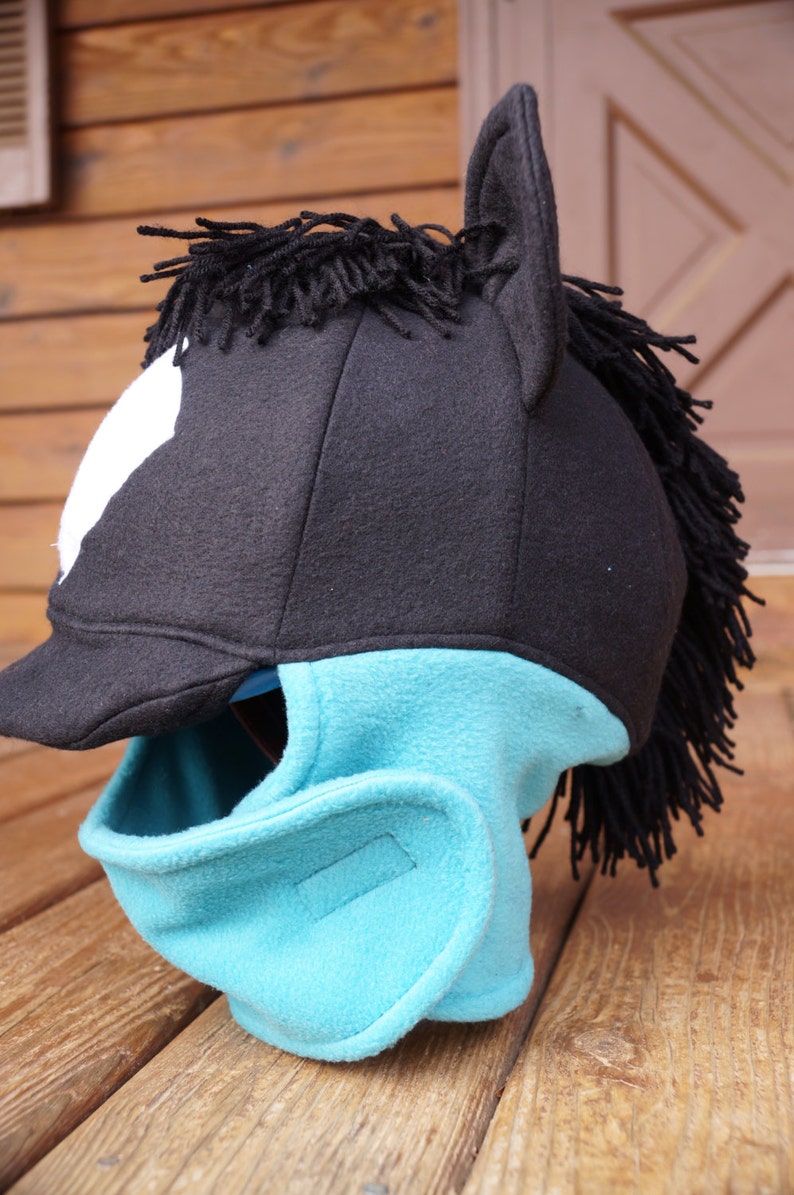 Puede incluir: Un gorro de caballo de lana negra y turquesa con una crin negra y una mancha blanca en la frente. El gorro tiene una visera y una m&aacute;scara facial.