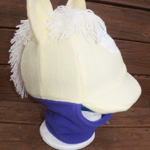 Puede incluir: Un gorro de cabeza de caballo de forro polar blanco y amarillo con un cuello de forro polar morado. El gorro tiene una crin blanca y una mancha blanca en la frente. El cuello tiene un agujero para la boca.