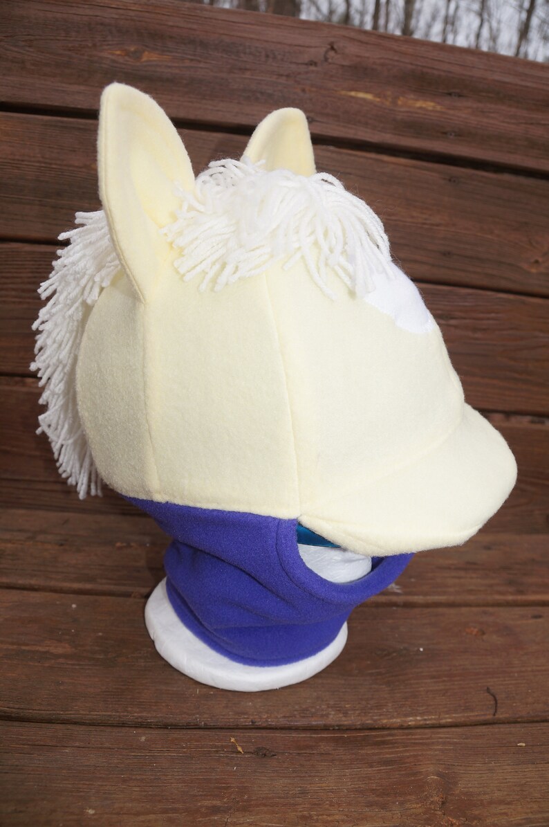 Puede incluir: Un gorro de cabeza de caballo de forro polar blanco y amarillo con un cuello de forro polar morado. El gorro tiene una crin blanca y una mancha blanca en la frente. El cuello tiene un agujero para la boca.