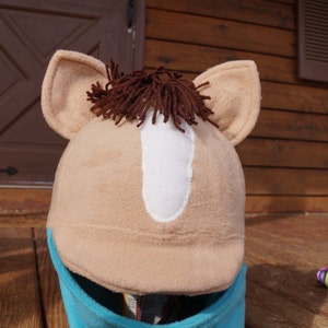 Puede incluir: Un casco de cabeza de caballo marr&oacute;n y blanco con un cuello de forro polar azul. El casco est&aacute; hecho de tela suave y tiene una crin marr&oacute;n.