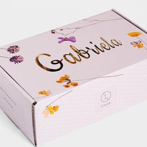 Pode incluir: Uma caixa de cart&atilde;o branca com um design floral e o nome "Gabriela" escrito em letras douradas. A caixa tem um pequeno logotipo com a letra "L" e a palavra "Liush" impressa nela.