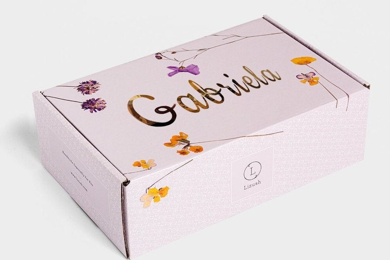 Pode incluir: Uma caixa de cart&atilde;o branca com um design floral e o nome "Gabriela" escrito em letras douradas. A caixa tem um pequeno logotipo com a letra "L" e a palavra "Liush" impressa nela.