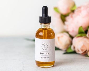 May include: A clear glass bottle with a black dropper top filled with a light amber liquid. The bottle has a white label with the text "Belly Oil" and a website address. The bottle is sitting on a white surface with a bouquet of pink flowers in the background.