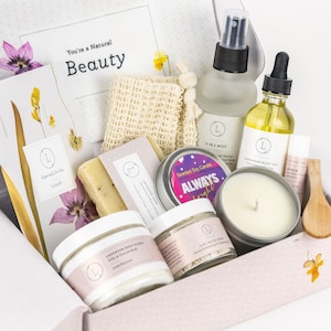 May include: A gift box filled with bath and body products, including a lavender body oil, a 2-in-1 mist, a soy candle, a bar of soap, a body scrub, and a facial clay mask. The box is decorated with dried flowers and the text "You're a Natural Beauty".
