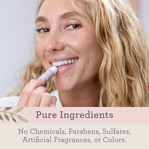 May include: A woman applies a lip balm to her lips. The lip balm tube is white with a pink label that says "Pure Ingredients". The text below the label says "No Chemicals, Parabens, Sulfates, Artificial Fragrances, or Colors."
