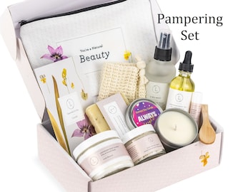 May include: A Pampering Set in a white box containing a zippered pouch, a spray bottle, a dropper bottle, a soy candle, a lip balm, a wooden spoon, a soap bar, a body scrub, a facial mask, and a textured washcloth with floral printed cards.